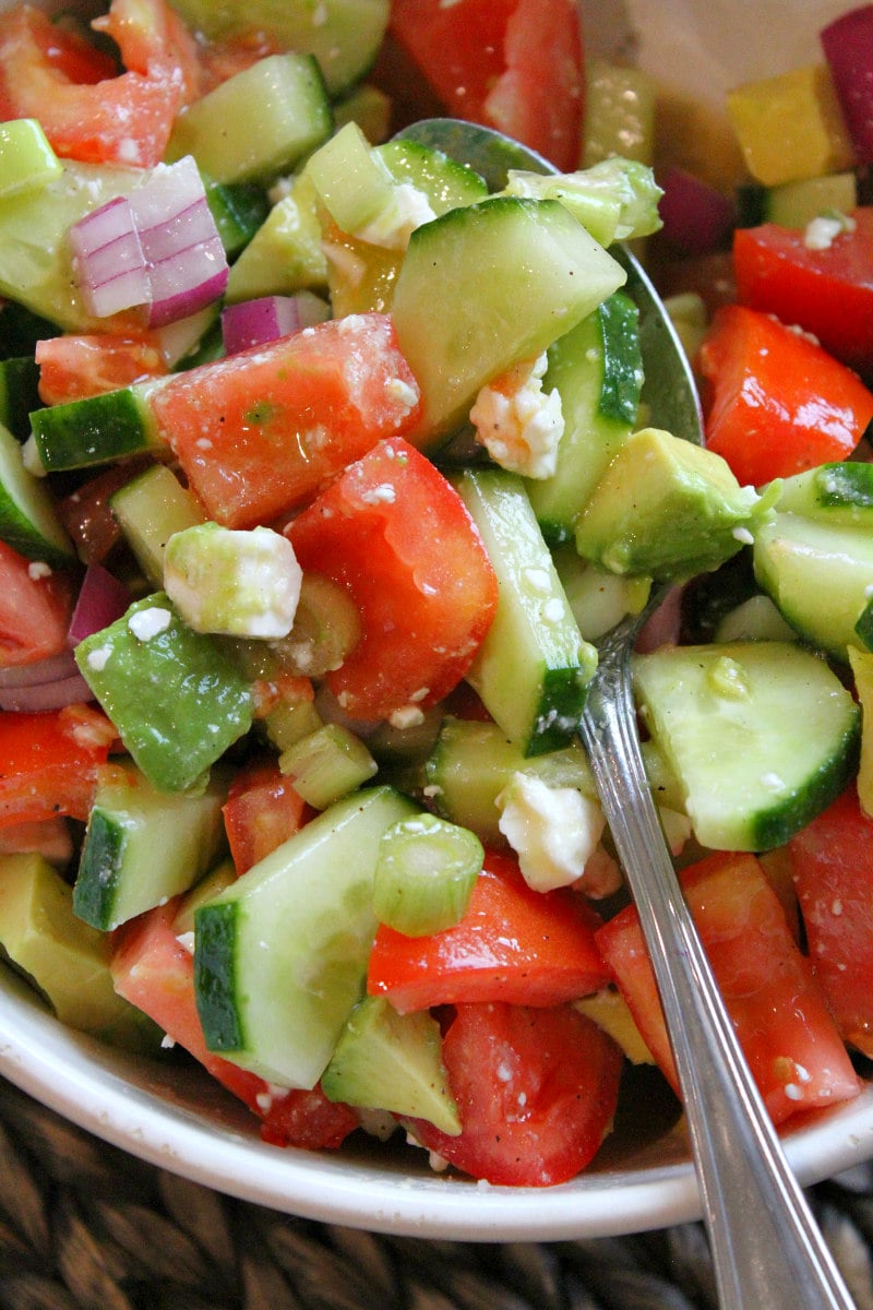 close up shot of avocado cucumber tomato salad in a white bowl with a serving spoon in it