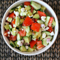 Overhead shot of avocado cucumber tomato salad in a white bowl placed on a dark brown woven place mat