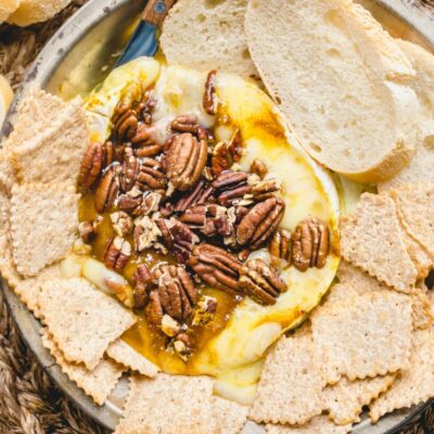 melted brie cheese with pecans on a platter surrounded by sliced baguette and crackers