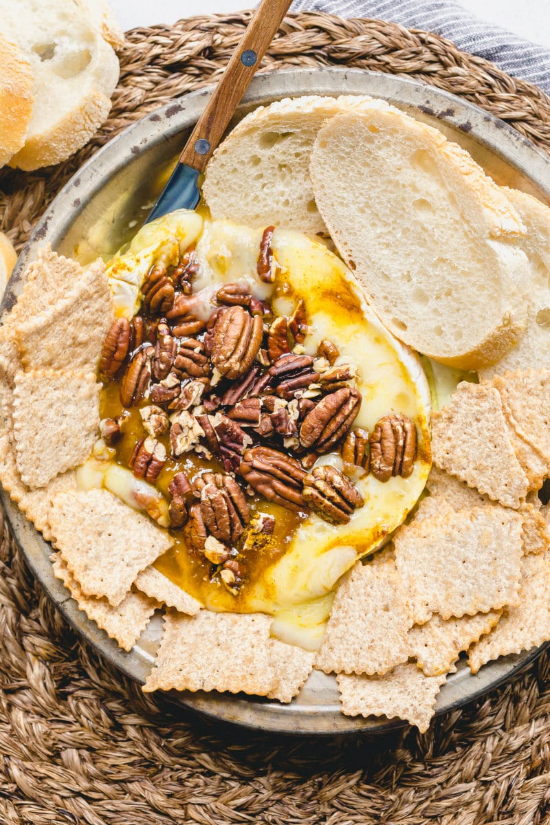 melted brie cheese with pecans on a platter surrounded by sliced baguette and crackers