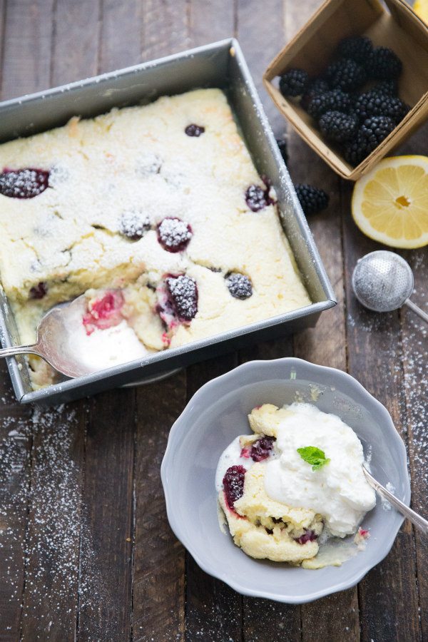 Serving blackberry Lemon Pudding Cake in a gray bowl with cream and a spoon. the rest of the pan of cake in the background with fresh blackberries and lemon displayed too