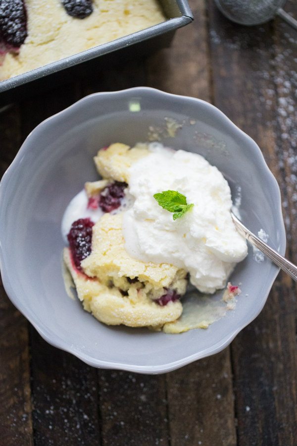 serving of Blackberry Lemon Pudding Cake with cream in a gray bowl. peek at the pan of cake in the background