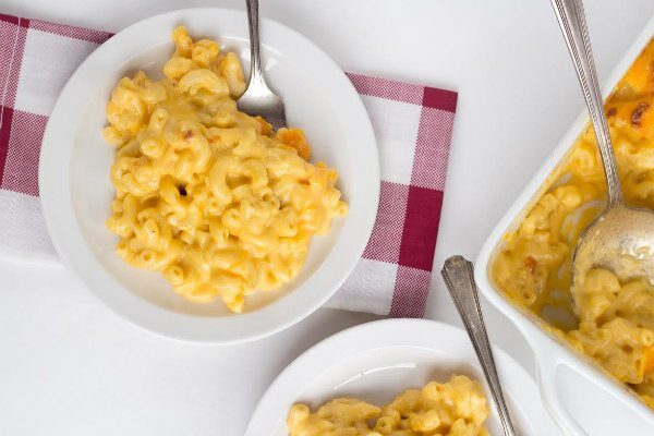 Overhead shot of macaroni and cheese on a white plate. Plaid red and white napkin underneath. Fork on plate. Peek at another plated mac and cheese and casserole dish of mac and cheese in background.
