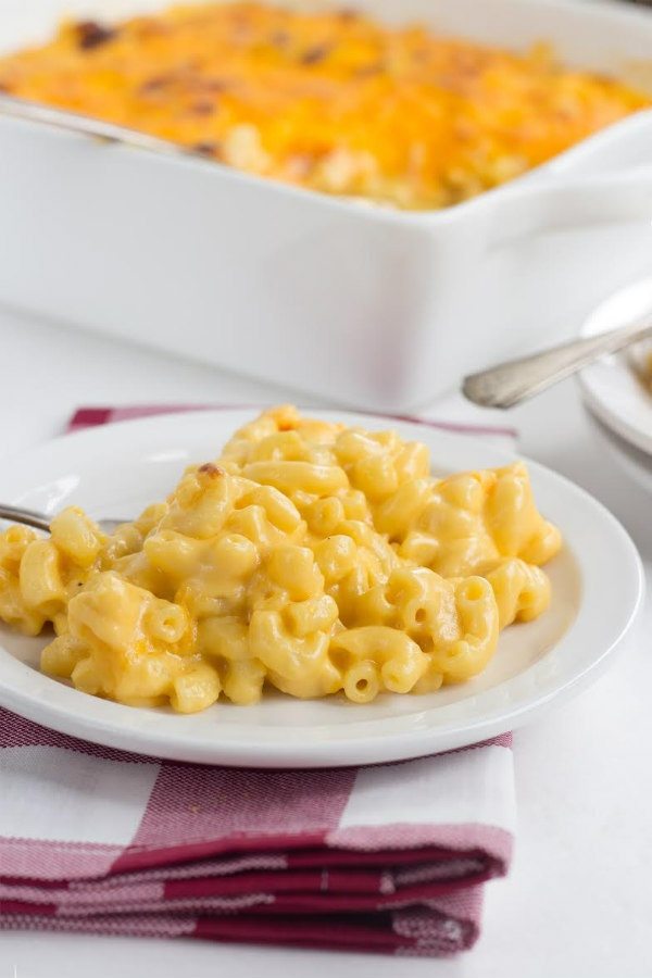 macaroni and cheese on a white plate with a red/white plaid napkin underneath and a white casserole dish in the background