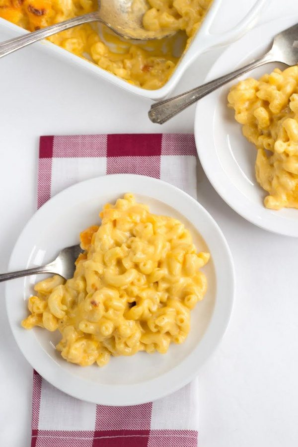 Overhead shot of macaroni and cheese on a white plate. Plaid red and white napkin underneath. Fork on plate. Peek at another plated mac and cheese and casserole dish of mac and cheese in background.