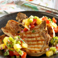 Grilled Pork Chops with tropical salsa on top set on a black dinner plate and a flowered napkin in the background