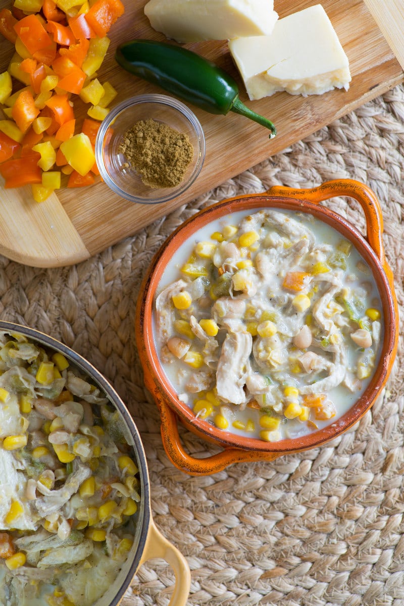 overhead shot of Bowl of White Chicken Chili in an orange bowl with pot of chili in background and cutting board with ingredients on top. set on top of woven placemat
