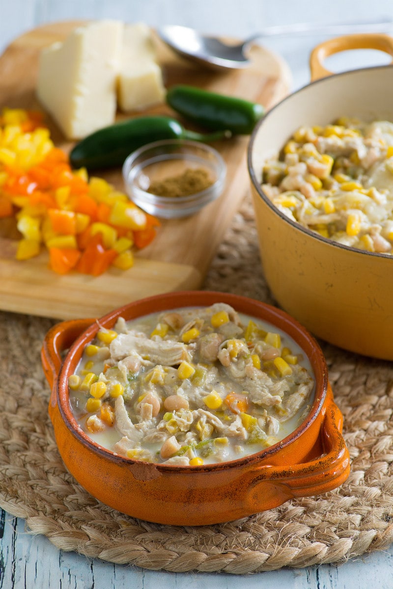 Bowl of White Chicken Chili in an orange bowl on a woven placemat with pot of chili in the background and cutting board with cut veggies and cheese on it