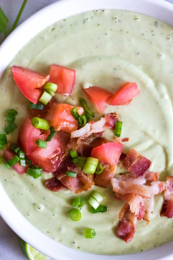 overhead shot of a Bowl of Avocado Soup topped with tomato and bacon and green onions- in a white bowl