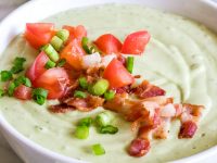chilled avocado soup in a white bowl topped with green onion and tomato. lime wedges, cilantro and another bowl of soup in the background