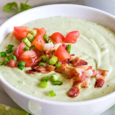 chilled avocado soup in a white bowl topped with green onion and tomato. lime wedges, cilantro and another bowl of soup in the background