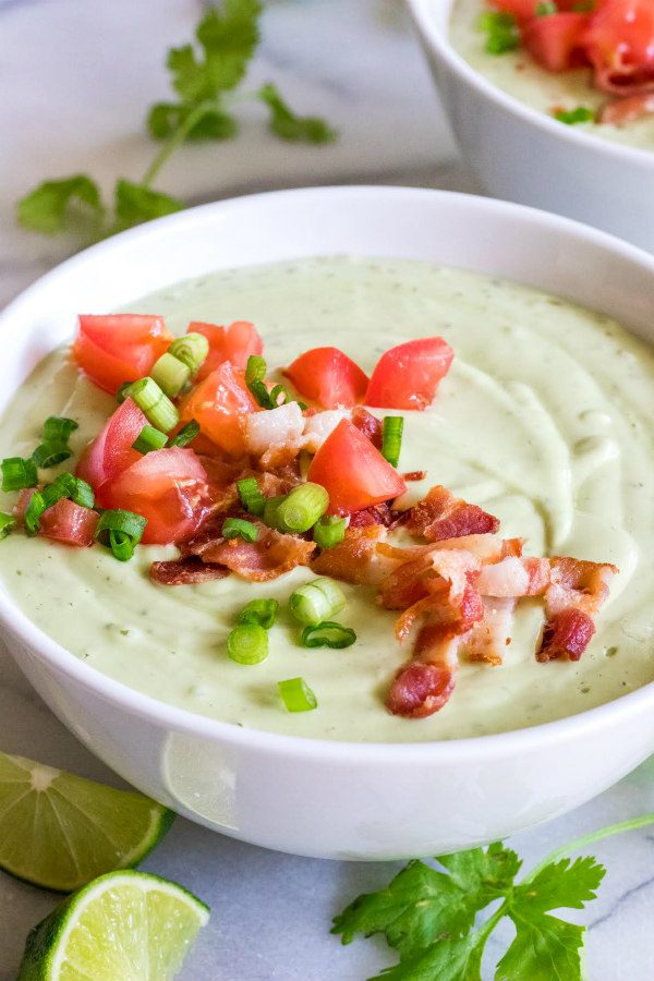 Bowl of Chilled Avocado Soup in a white bowl topped with tomato and bacon and green onions. Cilantro, lime wedges and another bowl of soup in the background