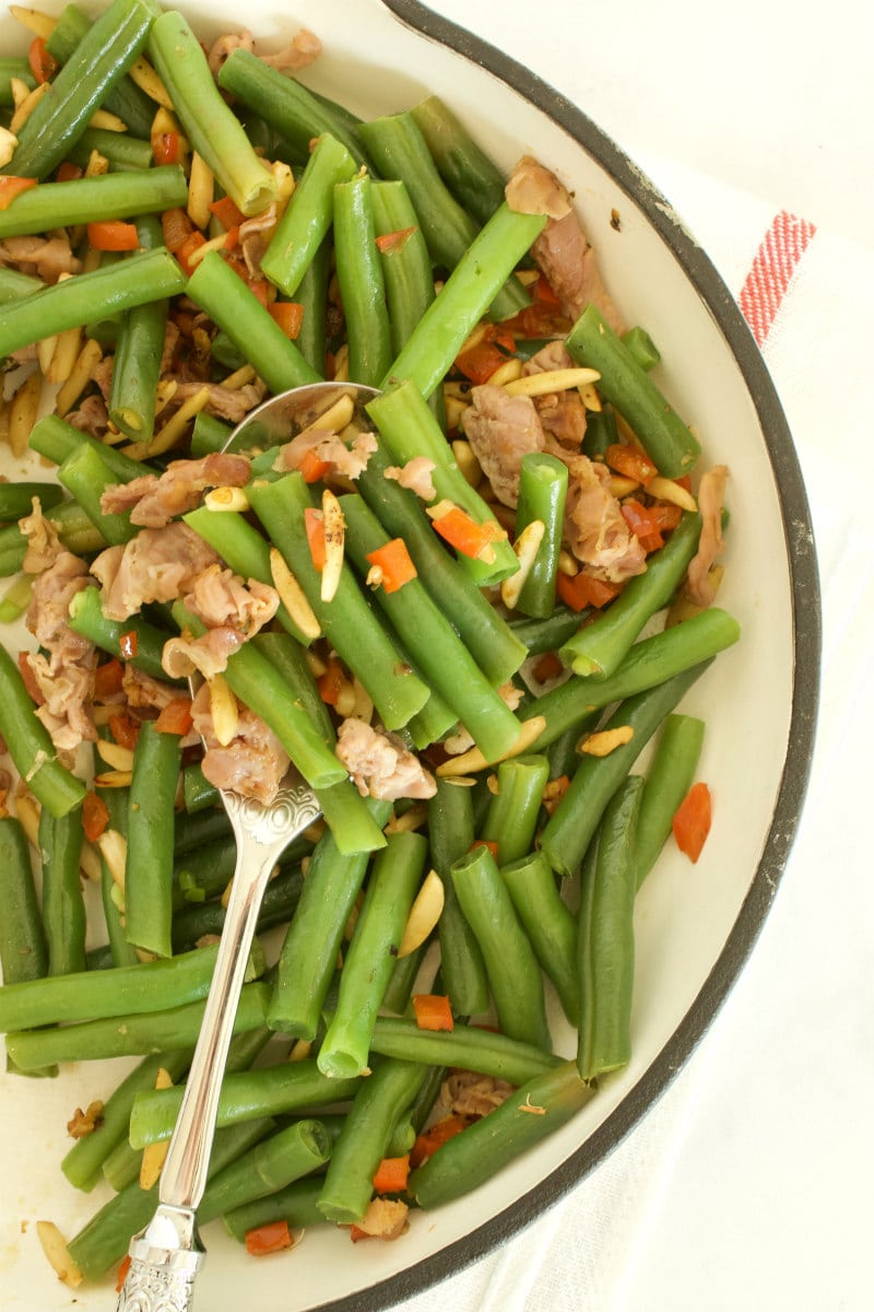 overhead shot of green beans with prosciutto in a fry pan with a serving spoon, set on a yellow napkin with red stripes