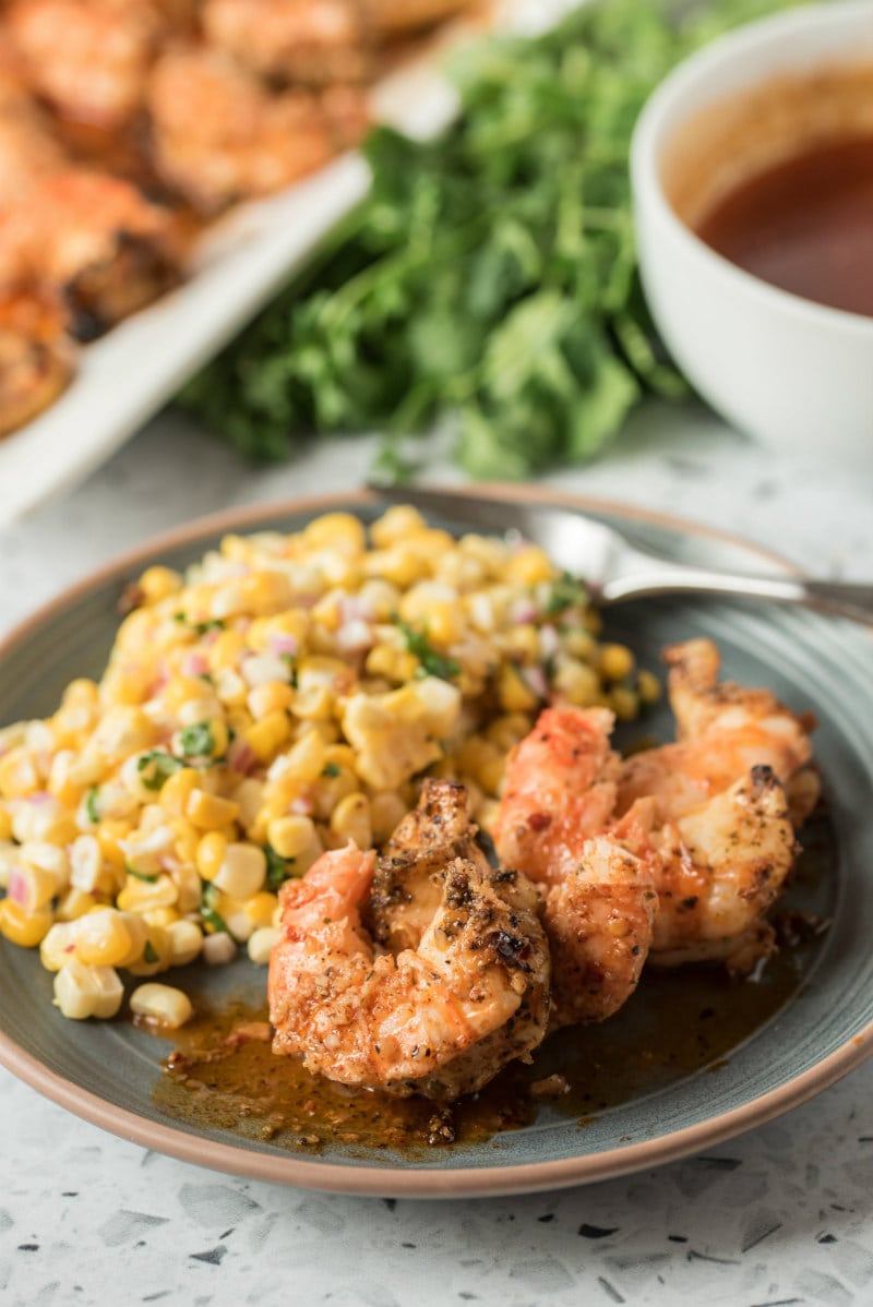 dinner plate with grilled jumbo shrimp and corn salad on it. lemon chipotle butter in a bowl displayed on the side with fresh cilantro and a tray of more shrimp in the background