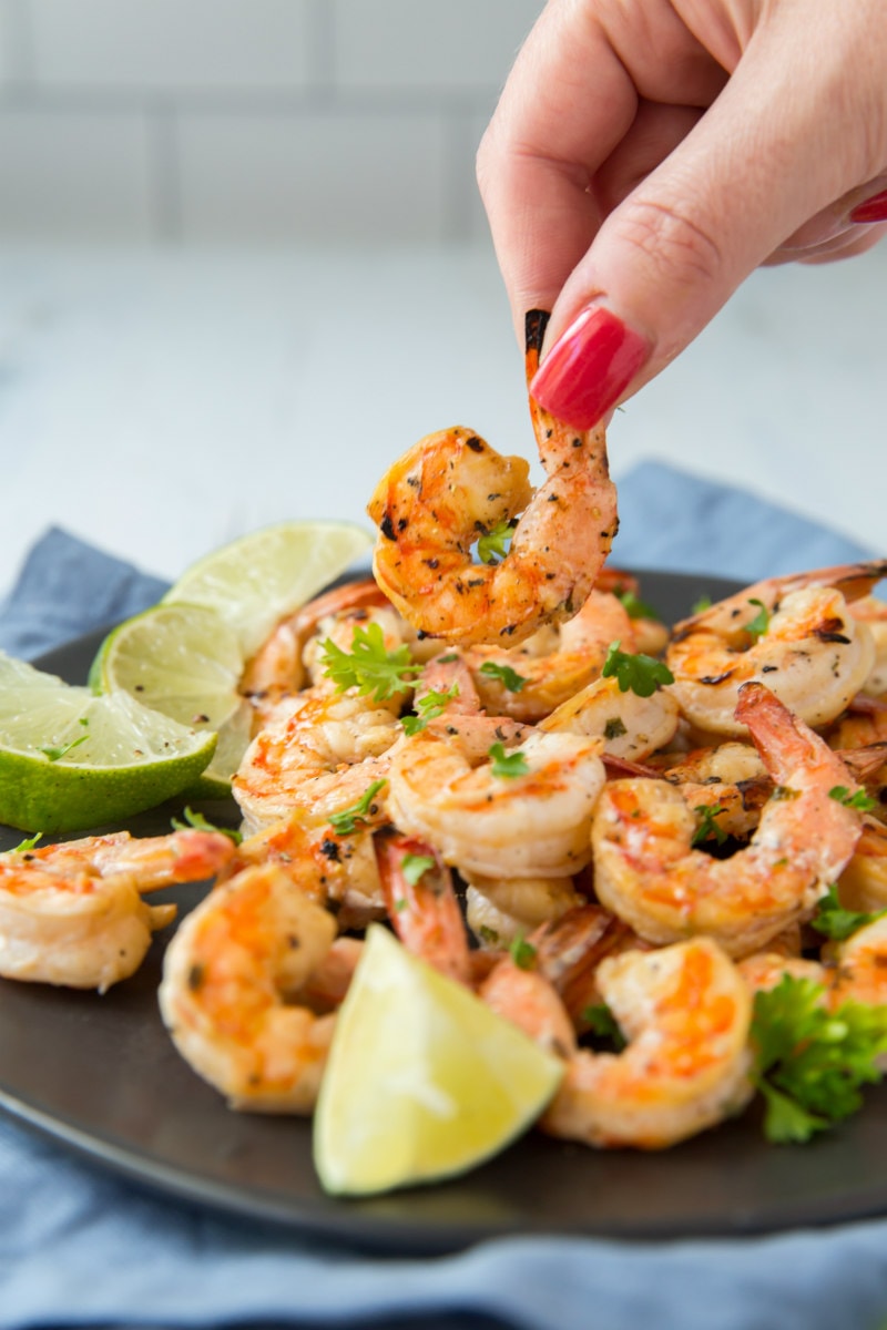 picking up a grilled shrimp off a black platter of shrimp with lime garnish and a blue napkin underneath