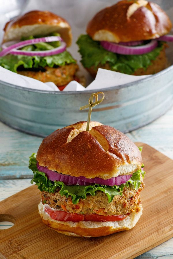 Turkey Garden Burger dressed with red onion, lettuce and tomato on a bun sitting on a wooden cutting board. galvanized metal tub in the background with two more burgers inside