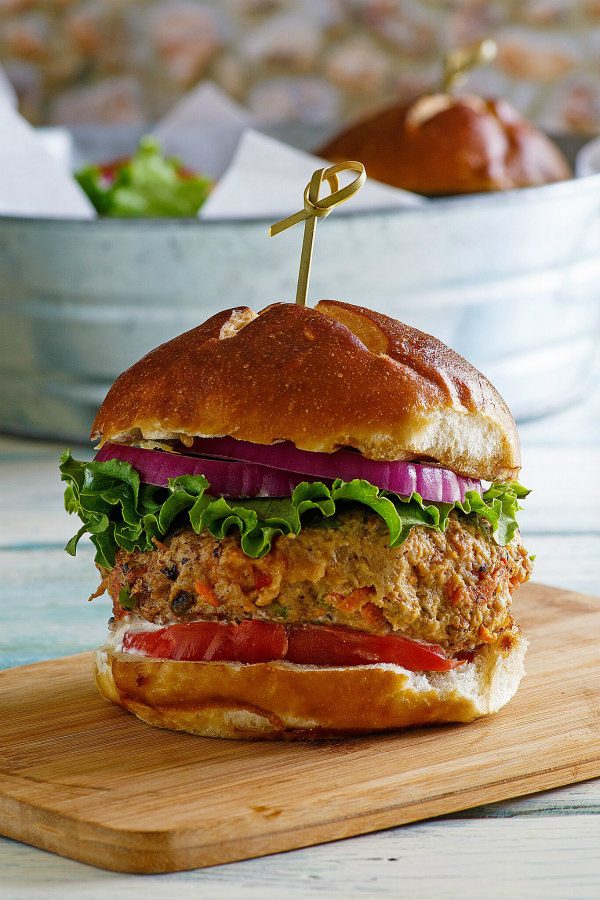 Turkey Garden Burger dressed with red onion, lettuce and tomato on a bun sitting on a wooden cutting board. galvanized metal tub in the background