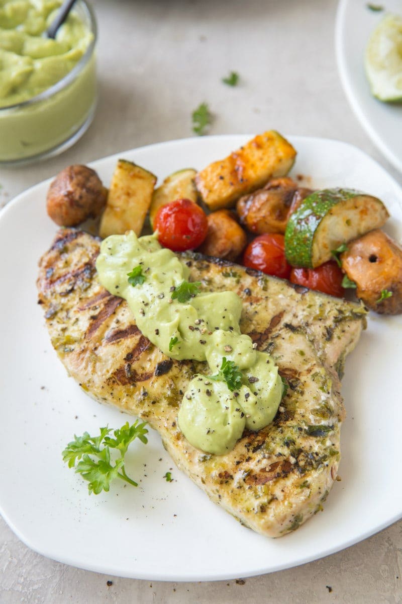 grilled swordfish topped with avocado mayonnaise on a white plate. Vegetables on the side.