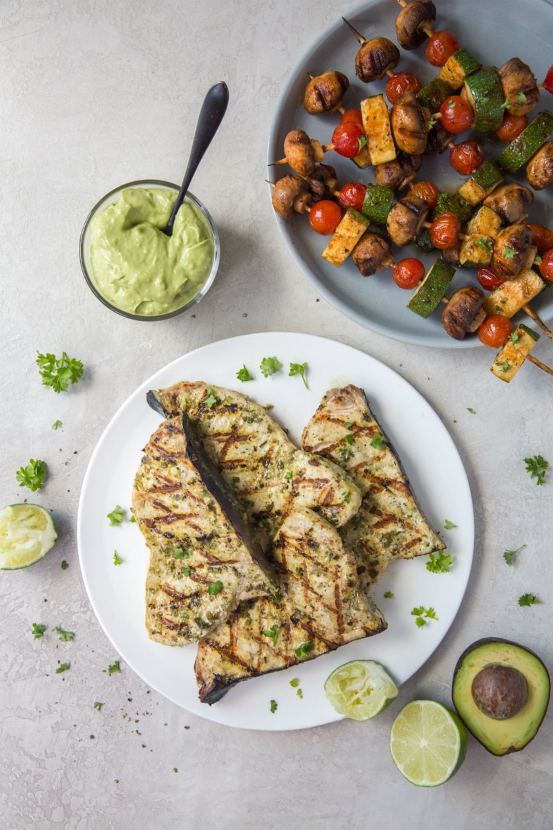 grilled swordfish on a white plate. Avocados and avocado mayonnaise displayed on the side. Vegetable kabobs displayed on the side.