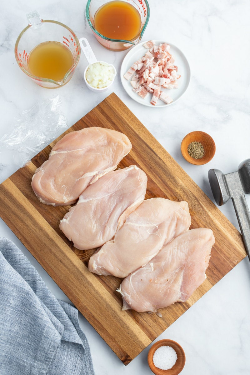 chicken on a cutting board with ingredients for making cider and bacon sauce displayed in bowls