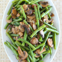 overhead shot of green beans with mushrooms and shallots on a white platter set on a woven white placemat