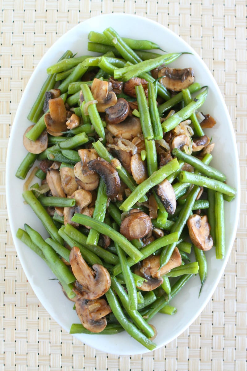 overhead shot of Green Beans with Mushrooms and Shallots on a white serving platter set on a woven white placemat