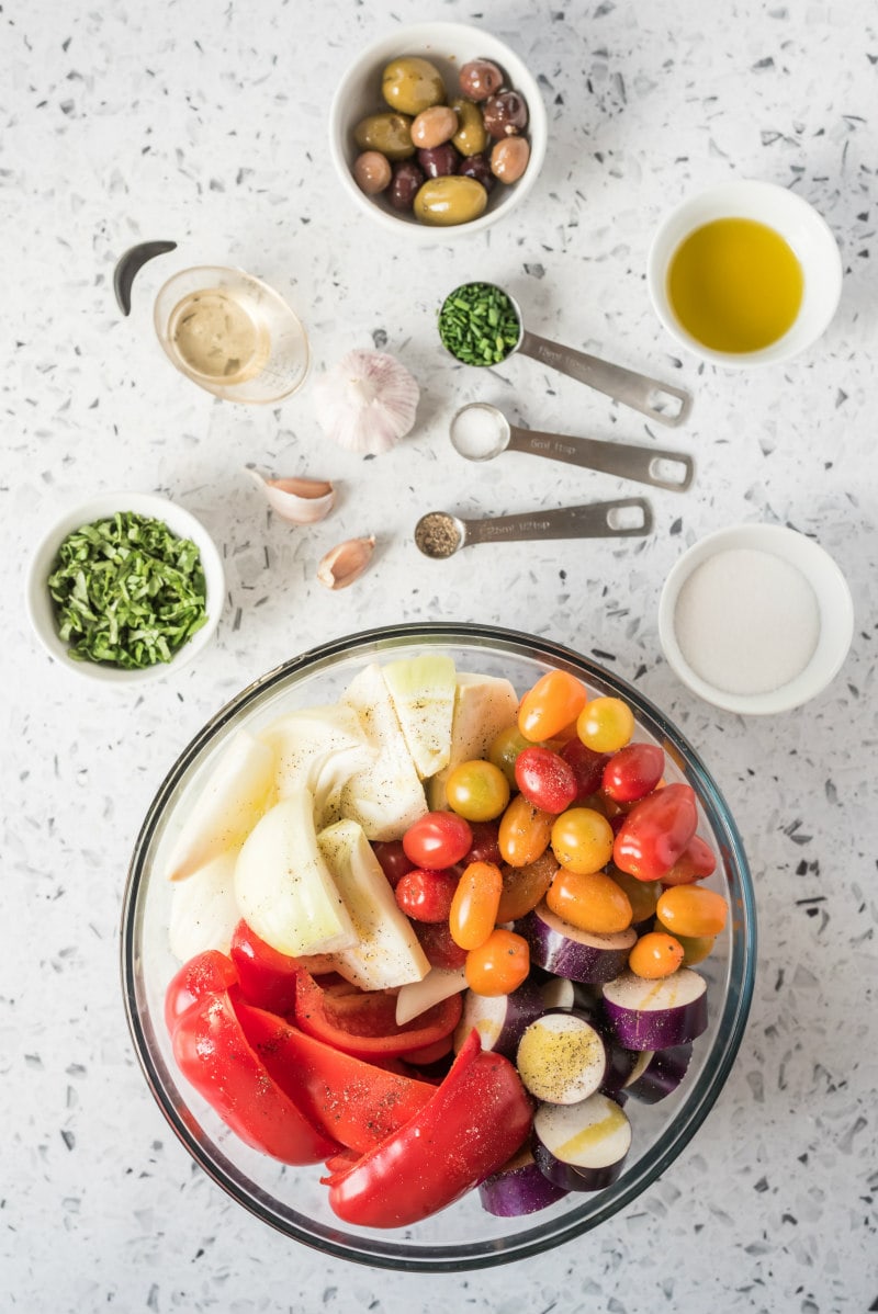 overhead shot of ingredients for grilled vegetable salad. glass bowl filled with vegetables- tomato, onion, red bell pepper, eggplant. measuring spoons and measuring cups with spices and other ingredients