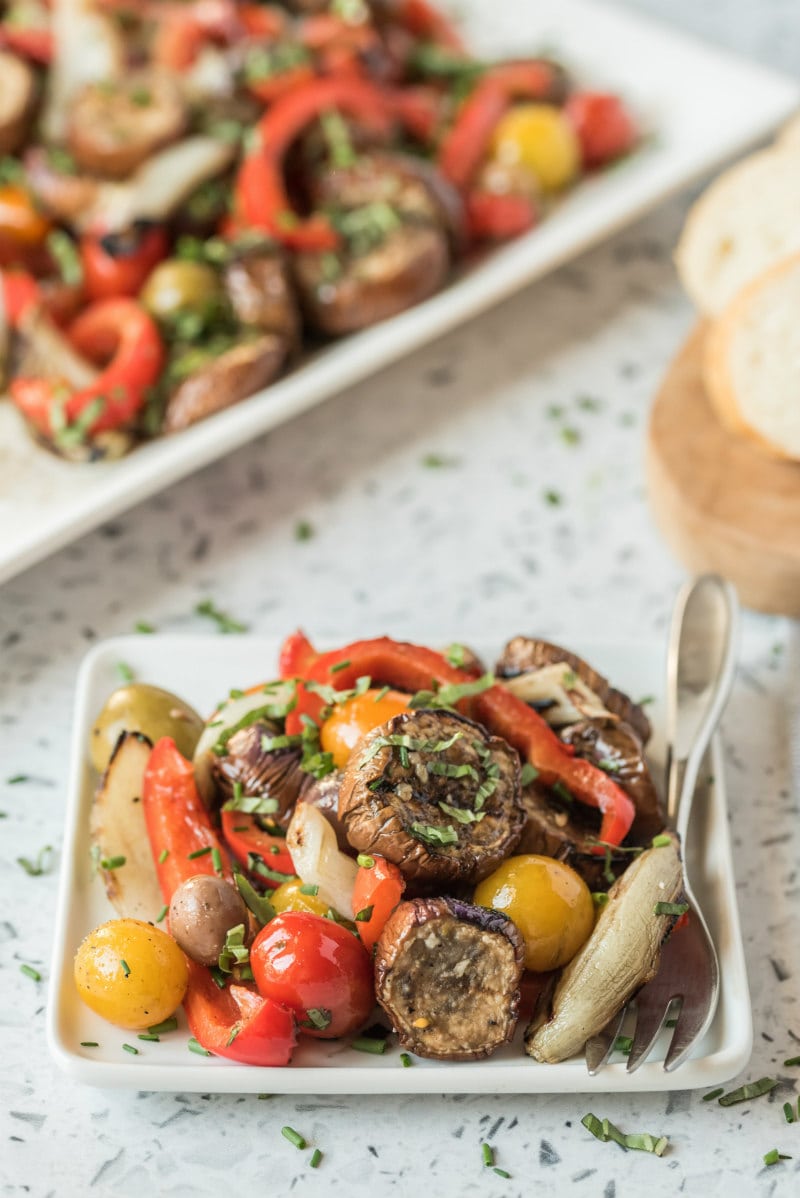 serving of grilled vegetable salad on a square white plate with the rest of the salad in the background. peek of bread slice on the side. fork on the plate.