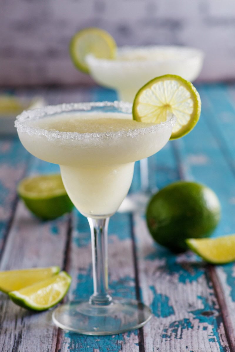 mock margarita in a margarita glass edged with salt and garnished with a lime slice. More limes and another margarita in the background. Set on a rustic turquoise board.