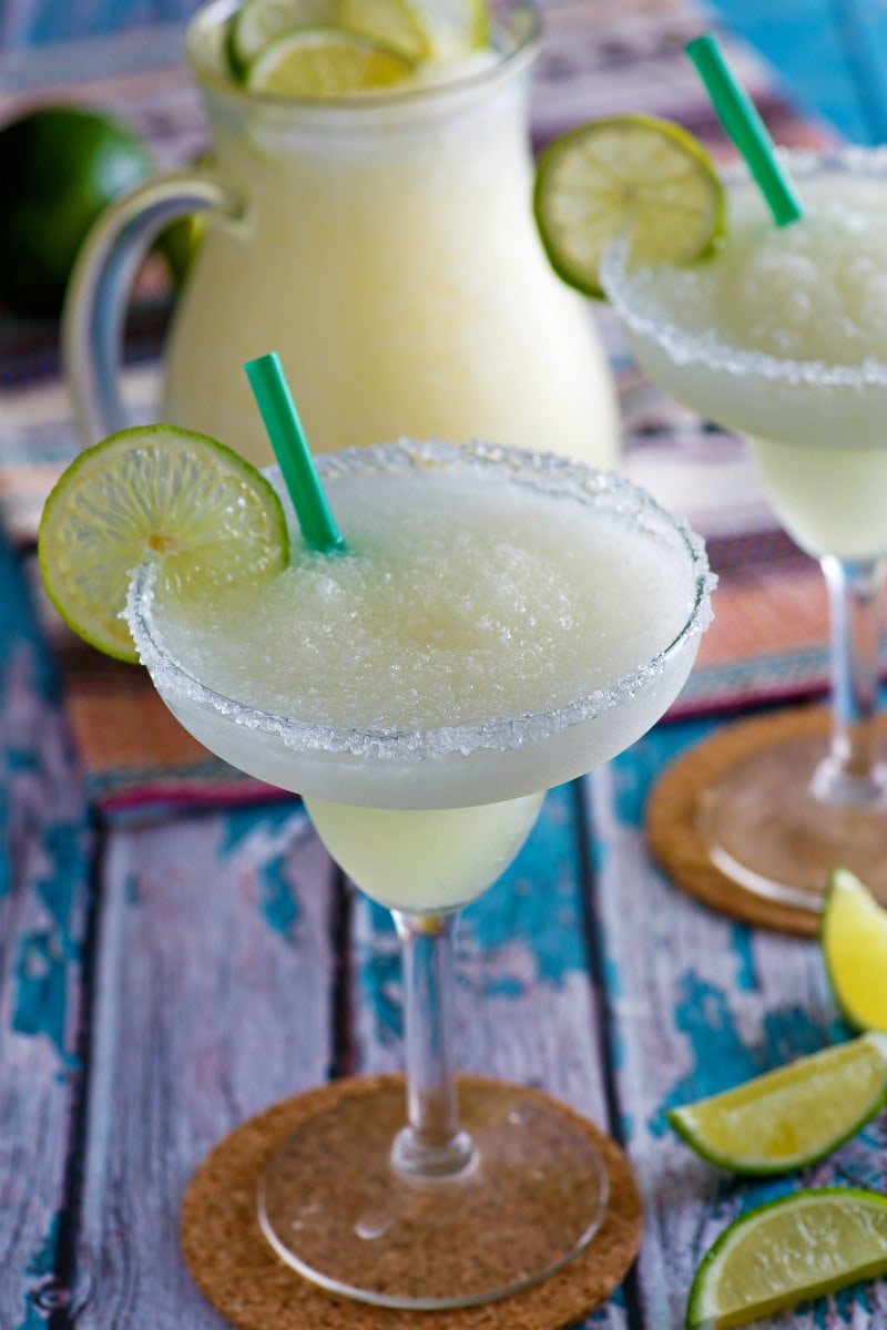 mock margarita in a margarita glass with a wine wedge and a straw. Pitcher of margaritas in background