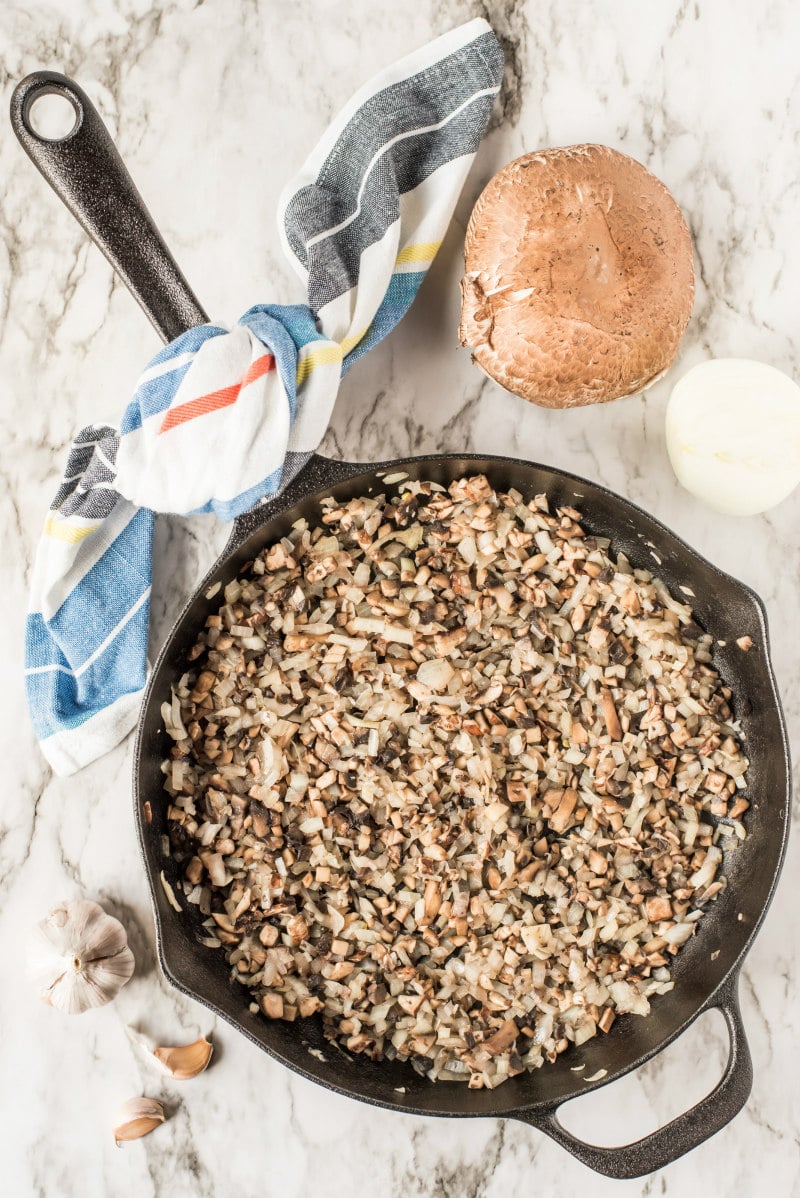 overhead shot of skillet full of cooked mushroom and onion. portobello mushroom and onion on the counter next to it. kitchen towel wrapped around the handle of the skillet