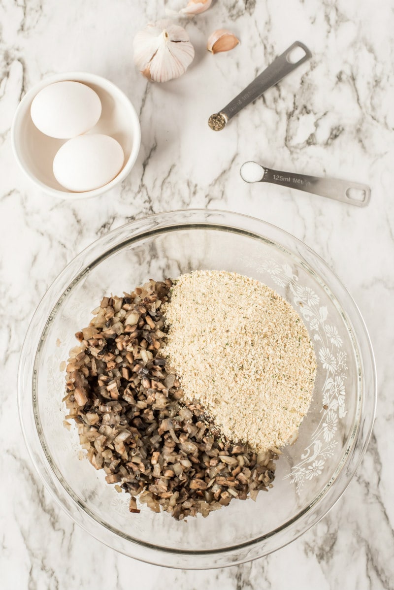 glass bowl with cooked mushroom and bread crumbs. two eggs in a white bowl on the counter next to it. measuring spoons on the counter too