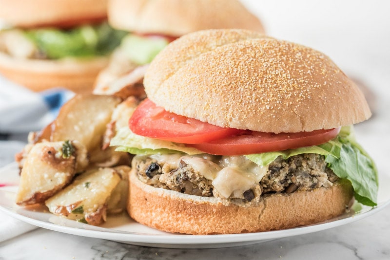 Mushroom Veggie Burgers recipe from RecipeGirl.com #mushroom #veggie #burgers #recipe #RecipeGirl mushroom veggie burger on a white plate garnished with lettuce and tomato. potato salad on the plate. more burgers in the background