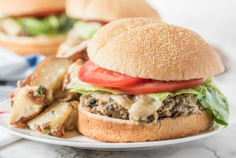 mushroom veggie burger on a white plate garnished with lettuce and tomato. potato salad on the plate. more burgers in the background