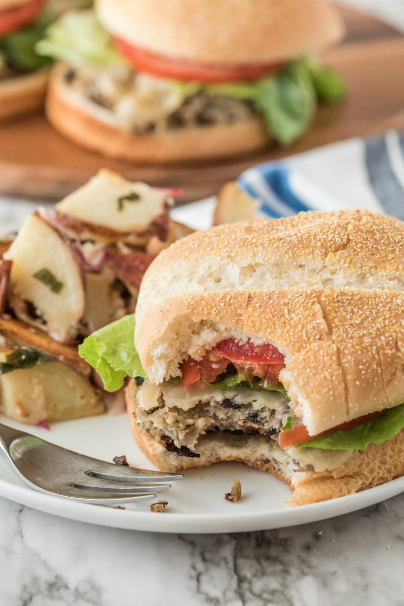 Mushroom Veggie Burgers recipe from RecipeGirl.com #mushroom #veggie #burgers #recipe #RecipeGirl mushroom veggie burger on a white plate with a bite taken out of it. fork and potato salad also on the plate. another burger in the background