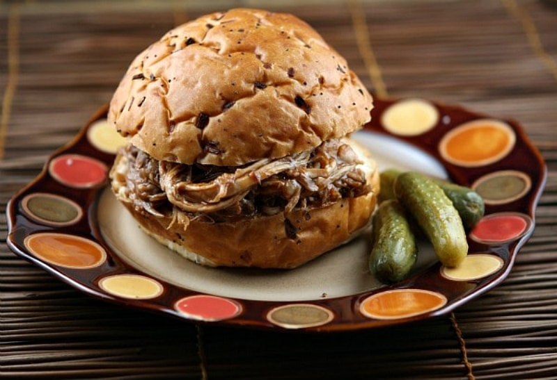 root beer pulled pork sandwich sitting on a brown and white plate with two pickles on the side on an orange and yellow polka dots