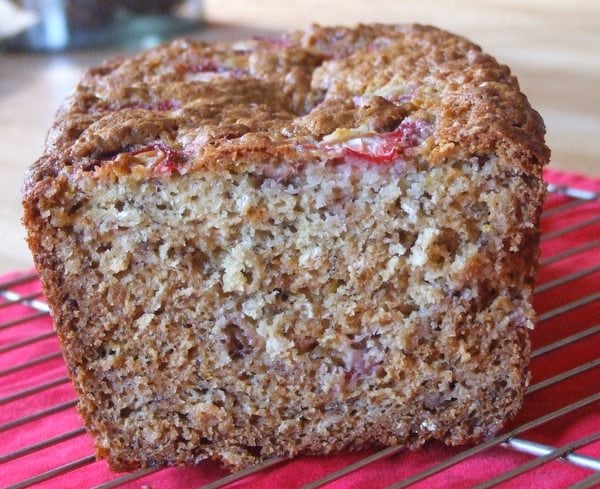 loaf of strawberry oatmeal banana bread sliced open to see the inside- sitting on a cooling rack on a red cloth napkin