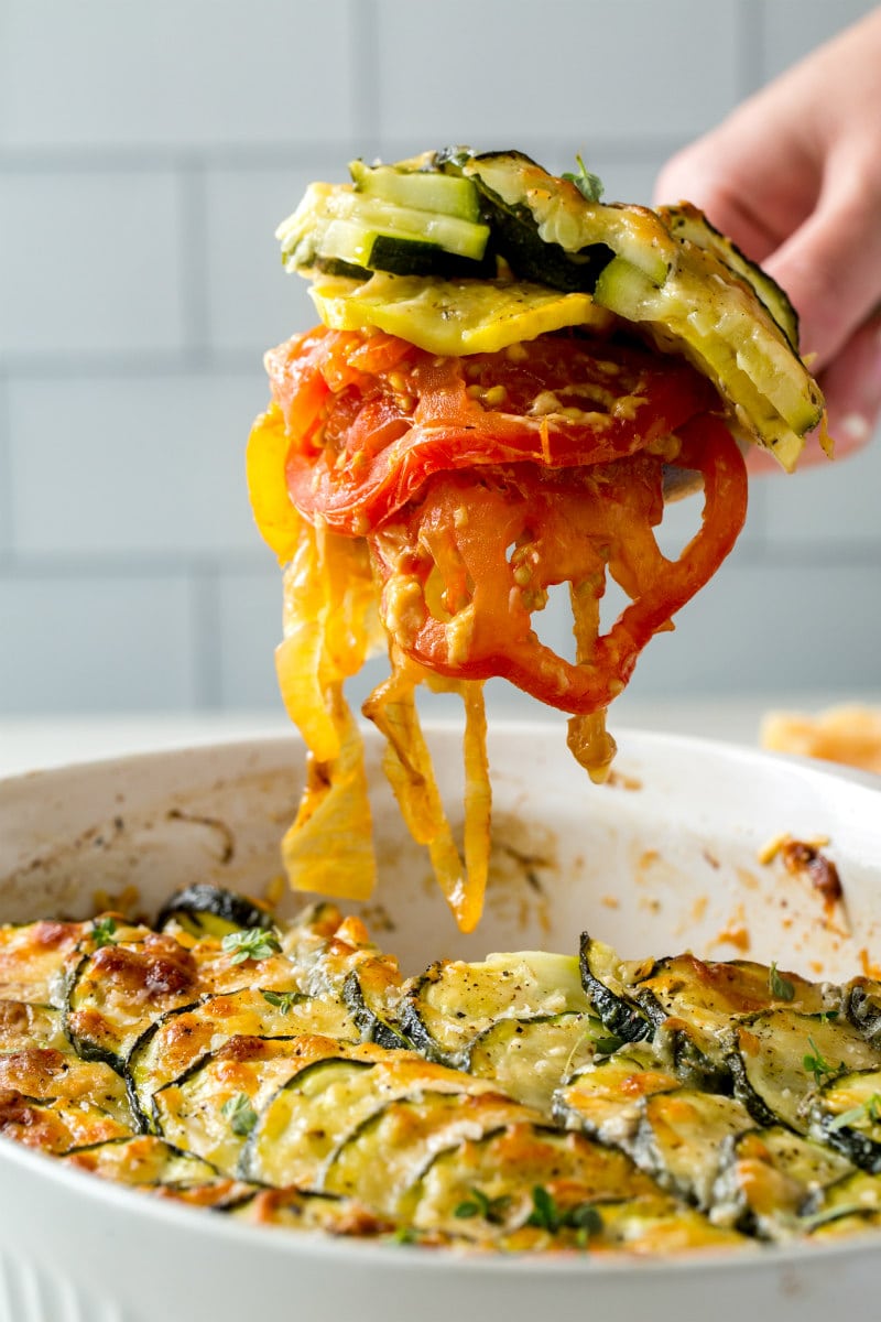 hand serving a spoonful of squash and tomato gratin over a casserole dish of gratin