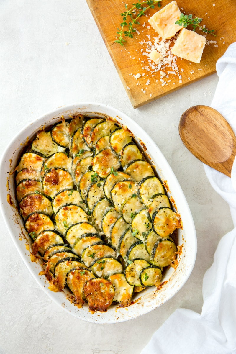 zucchini and tomato gratin in a white casserole dish with a wooden cutting board with cheese on it and a wooden spoon on the side