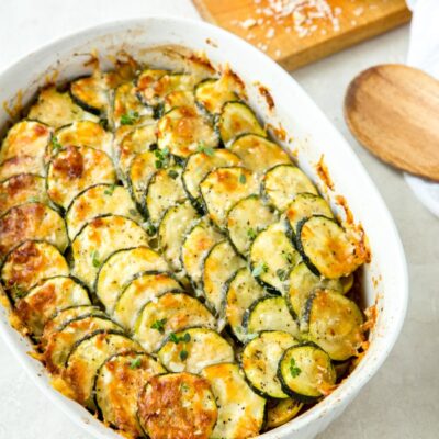 zucchini and tomato gratin in a white casserole dish with a wooden cutting board with cheese on it and a wooden spoon on the side