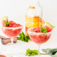 two watermelon mint margaritas garnished with mint and watermelon with a bottle of tequila in the background