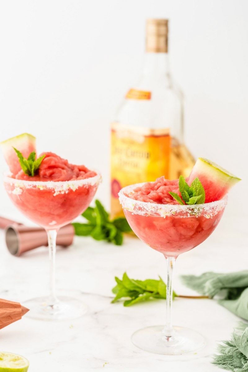 two watermelon mint margaritas garnished with mint and watermelon with a bottle of tequila in the background