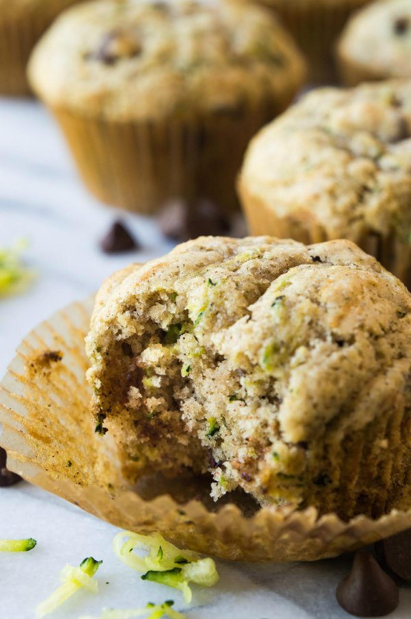 zucchini chocolate chip muffin with a bite out of it sitting on a marble surface with chocolate chips and shredded zucchini scattered about and more muffins in the background