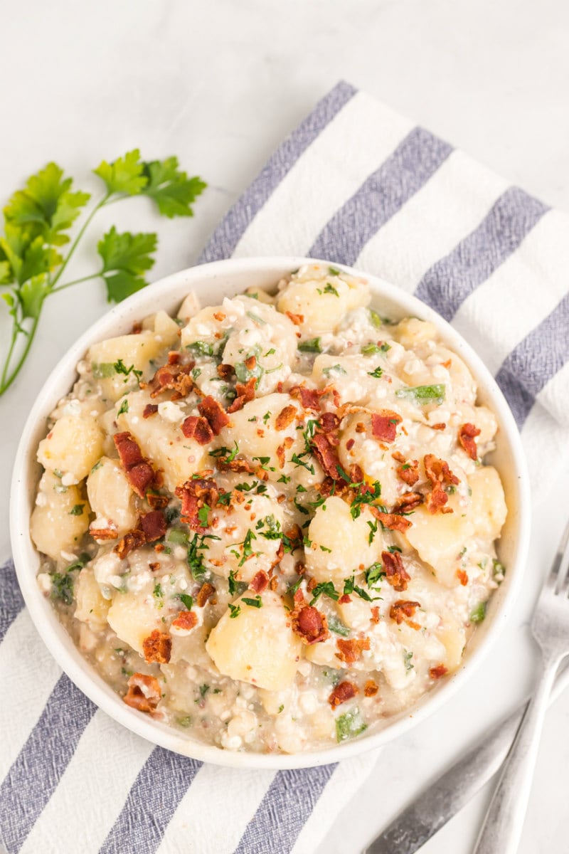 blue cheese and bacon potato salad in a white bowl with blue and white striped cloth napkin with parsley garnish