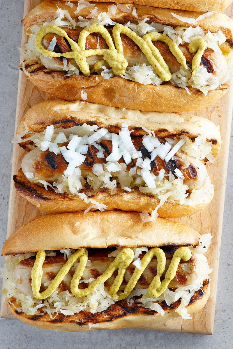 overhead shot of three bratwurst subs on a wooden board topped with mustard, onion and/or sauerkraut