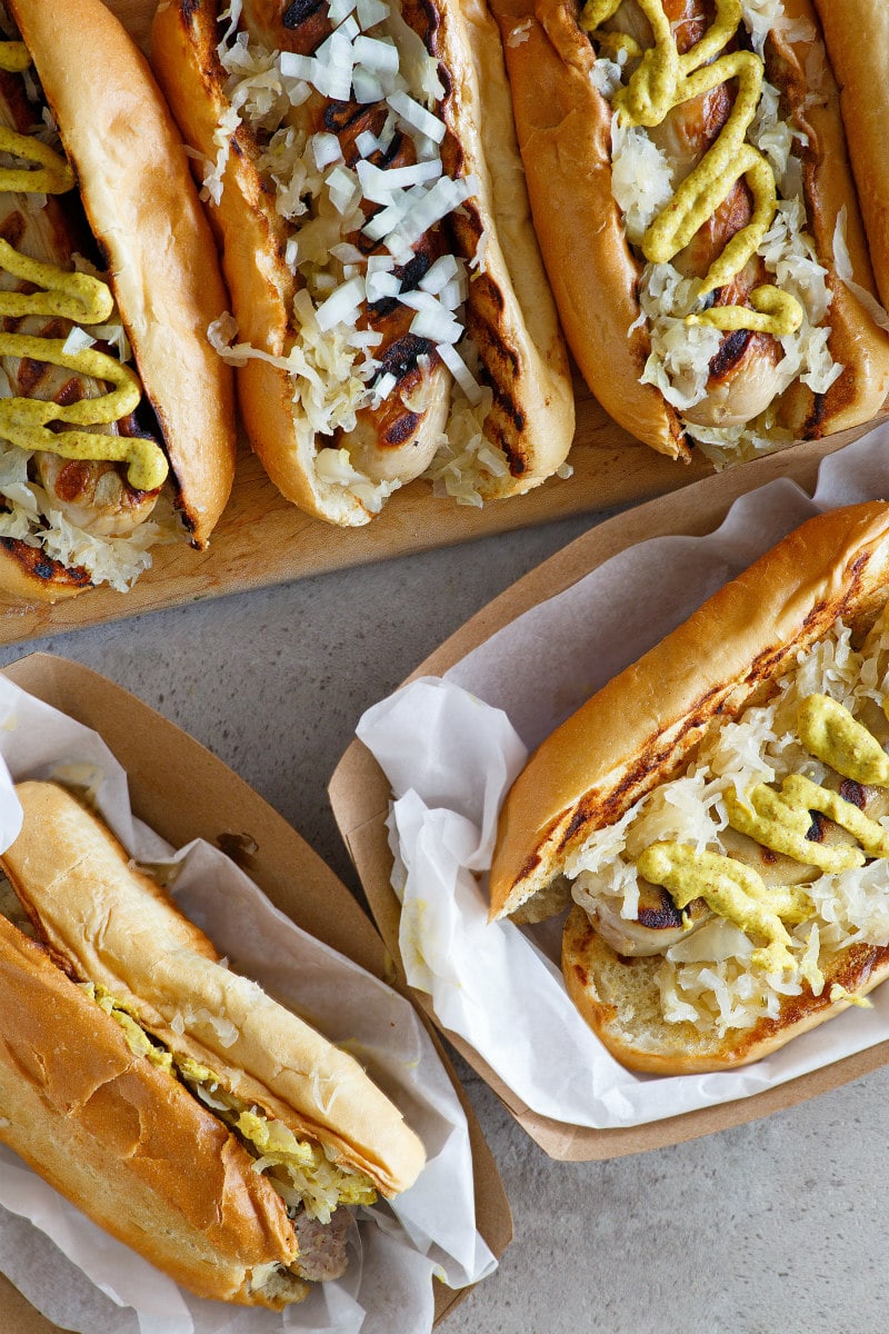 2 Bratwurst Subs displayed on serving trays and 3 more subs on a wooden board in the background. the subs are topped with mustard, onion and/or sauerkraut