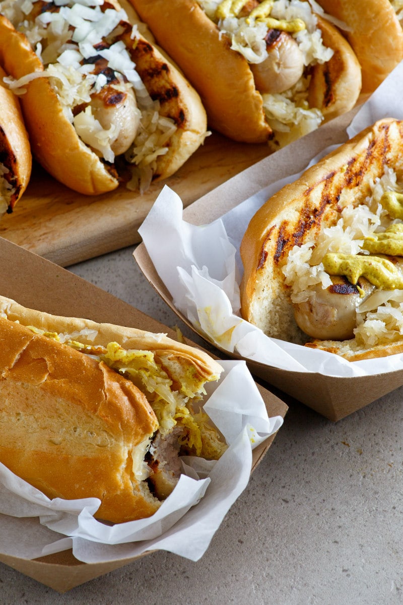 2 bratwurst subs in serving trays topped with mustard, onion, saurkraut. more bratwurst subs on a cutting board in the background