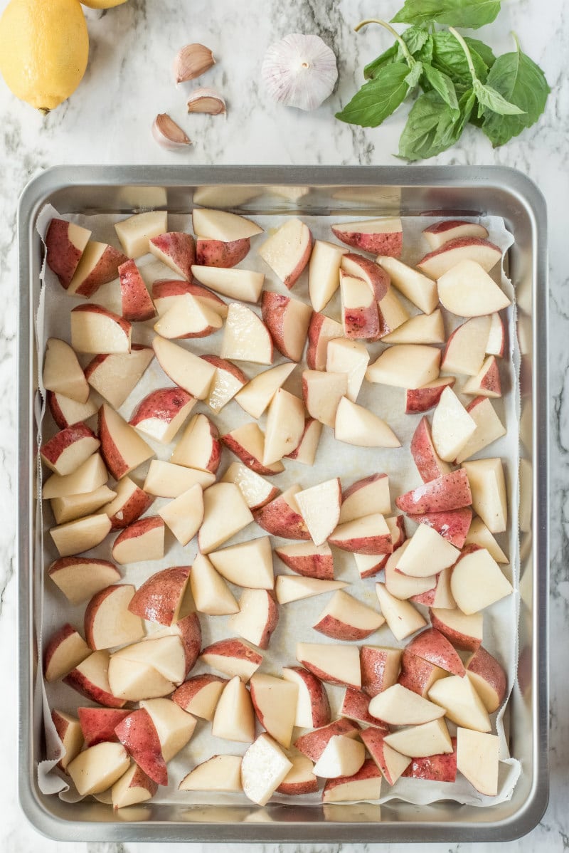 sheet pan filled with cut red potatoes ready for roasting and fresh basil, garlic and lemon on the side