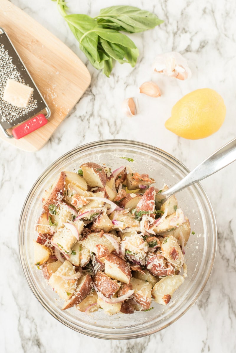 overhead shot of glass bowl of lemon basil roasted potato salad with lemon, garlic, basil and cutting board with cheese and shredder