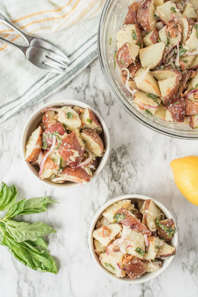 overhead shot of lemon basil roasted potato salad in a glass bowl with two individual servings in white bowls. striped napkin in view with forks, fresh basil and lemon